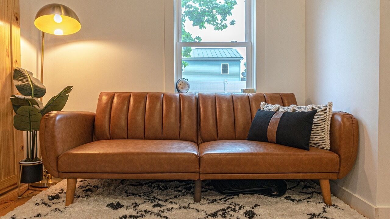 A brown leather sofa with a black and white patterned pillow, a floor lamp, and a plant in a corner.