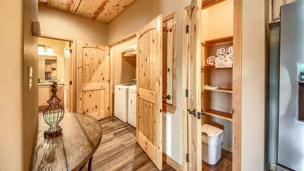 Un couloir avec des portes en bois, un lave-linge, un réfrigérateur et une table ornée d’un objet décoratif.