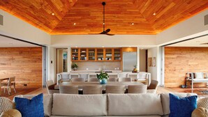 Uma sala de estar moderna com teto de madeira, um sofá grande e uma área de refeições com cozinha ao fundo.