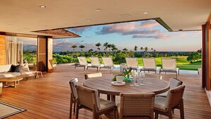 Um deck espaçoso ao ar livre com uma mesa redonda e cadeiras, com vista para uma paisagem exuberante com palmeiras e o oceano.