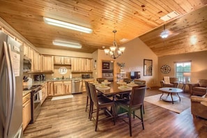 A spacious kitchen with wooden cabinets, a dining table with chairs, a chandelier, and a living area with sofas and a coffee table.