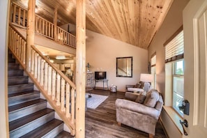 A wooden staircase leading to a second floor, a living room with a sofa and a TV, and a large window with blinds.