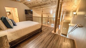 A bedroom with a wooden ceiling, a bed with white bedding, a wooden floor, a TV on a wooden cabinet, and a balcony with a wooden railing.