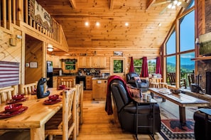 A wooden cabin interior with a dining area, kitchen, and large windows overlooking a forested landscape.