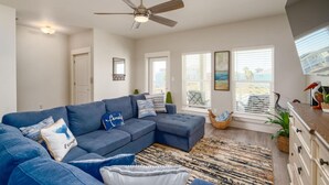 A spacious living room with a sectional sofa, a ceiling fan, and large windows offering a view of the outdoors. - Dive In and Unwind: Pet-Friendly Living with Community Pools and Hot Tubs. (Port Aransas)