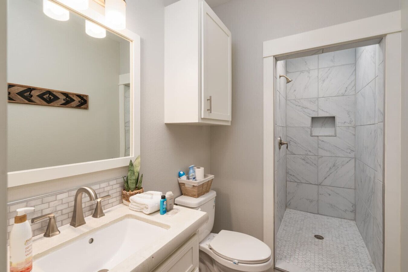 A bathroom with a white sink, a toilet, a shower with marble tiles, and a mirror with lights.
