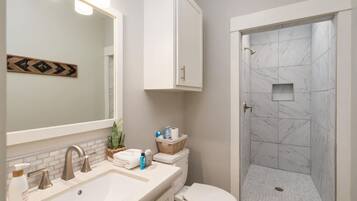 A bathroom with a white sink, a toilet, a shower with marble tiles, and a mirror with lights.