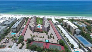 Vista aérea de um hotel com vários edifícios, uma piscina central, quadras de tênis e uma praia ao fundo.