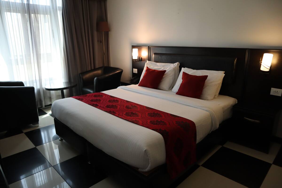 A hotel room with a checkered floor, a bed with white and red bedding, a nightstand with a lamp, and a chair.