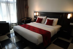 A hotel room with a checkered floor, a bed with white and red bedding, a nightstand with a lamp, and a chair.