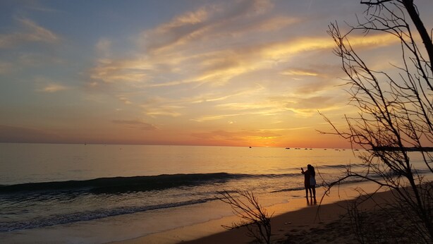 Une scène de plage avec un coucher de soleil, une mer calme et une personne debout sur le rivage.