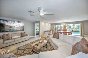 A spacious living room with a large sofa, a coffee table, and a dining area with a table and chairs. A ceiling fan is visible, and there is a large window with a view of the outdoors. - Walking Distance to the Beach! Canal / Pool Home! (Marco Island)