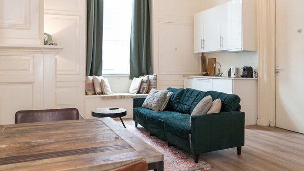 Une pièce comprenant une table à manger, des chaises, un canapé, une fenêtre avec des rideaux, un coin cuisine et un luminaire suspendu.
