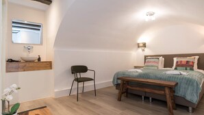 Une chambre à coucher avec un lit, une table de chevet, une chaise et une petite table sur laquelle est posé un vase de fleurs.