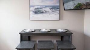 Un comedor con una mesa dispuesta para tres personas, un televisor de pared que muestra un paisaje marino y una zona de estar con banco.