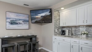 Una cocina con armarios blancos, salpicadero de azulejos, televisor de pantalla plana y una zona de bar con taburetes.