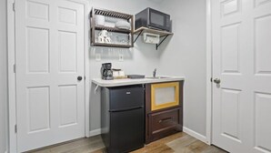 Una cocina compacta con puertas blancas, un refrigerador negro, un microondas y un estante con vajilla y vasos.