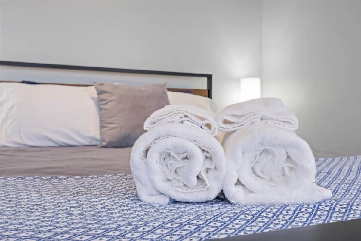 A neatly made bed with a patterned bedspread, a pillow, and rolled white towels on the floor.