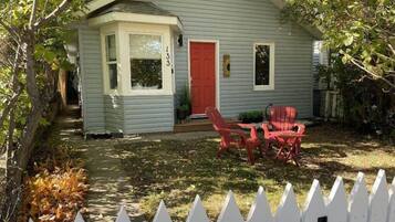 A small house with a red door, a white picket fence, and red chairs outside.