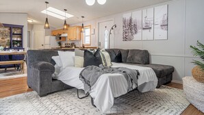 A modern living room with a grey sectional sofa, a white bed, and a dining area with wooden furniture. - Mountain Time | Furnished 30+ Day Retreat (Crowsnest Pass)
