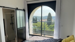 Un dormitorio con cama, ducha y un ventanal con vistas a zonas verdes y edificios.