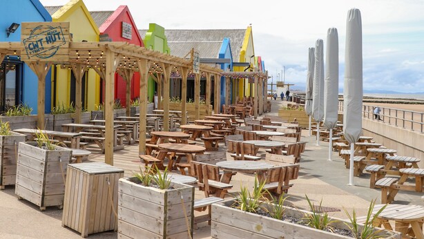 Ein Café am Strand mit Holztischen und -stühlen, bunten Gebäuden und Blick aufs Meer.