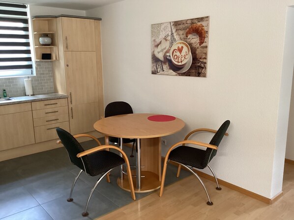 A dining area with a round table, four chairs, a red coaster, and a wall-mounted painting of a coffee cup. - Vacation apartment in the border triangle, 3 rooms 64 sqm, fully equipped. 1ST FLOOR (Steinen)