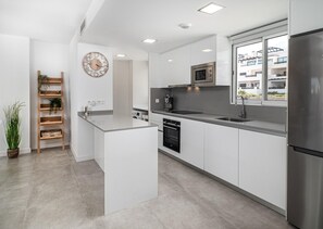 Une cuisine moderne avec des meubles blancs, un réfrigérateur en acier inoxydable, un four encastré et un micro-ondes. On y trouve une étagère en bois en forme d’échelle, ornée de plantes, une horloge au mur et une fenêtre donnant sur les bâtiments environnants.