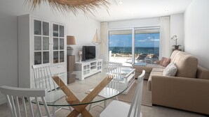 Un salón moderno con una mesa de comedor de vidrio, sillas blancas, un sofá color beige, un televisor sobre un mueble blanco y un ventanal con vistas al mar.