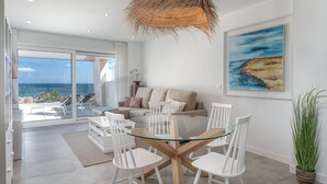 Una sala de estar moderna con una mesa de comedor de vidrio, sillas blancas, un sofá color beige y un cuadro con una playa.