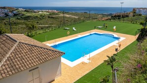 Une piscine avec vue sur la mer, entourée d’une pelouse bien entretenue et de quelques bâtiments en arrière-plan.