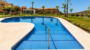 Ein Pool mit gewölbter Kante, umgeben von einer Betonplattform, Palmen und Gebäuden im Hintergrund.