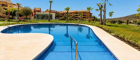 Une piscine à bord incurvé, entourée d’une terrasse en béton, de palmiers et de bâtiments en arrière-plan.