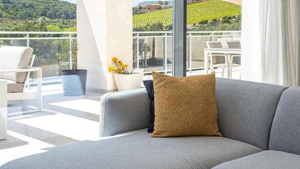 Ein modernes Wohnzimmer mit einem grauen Sofa, einem gemusterten Zierkissen und Blick auf einen Weinberg durch große Fenster.
