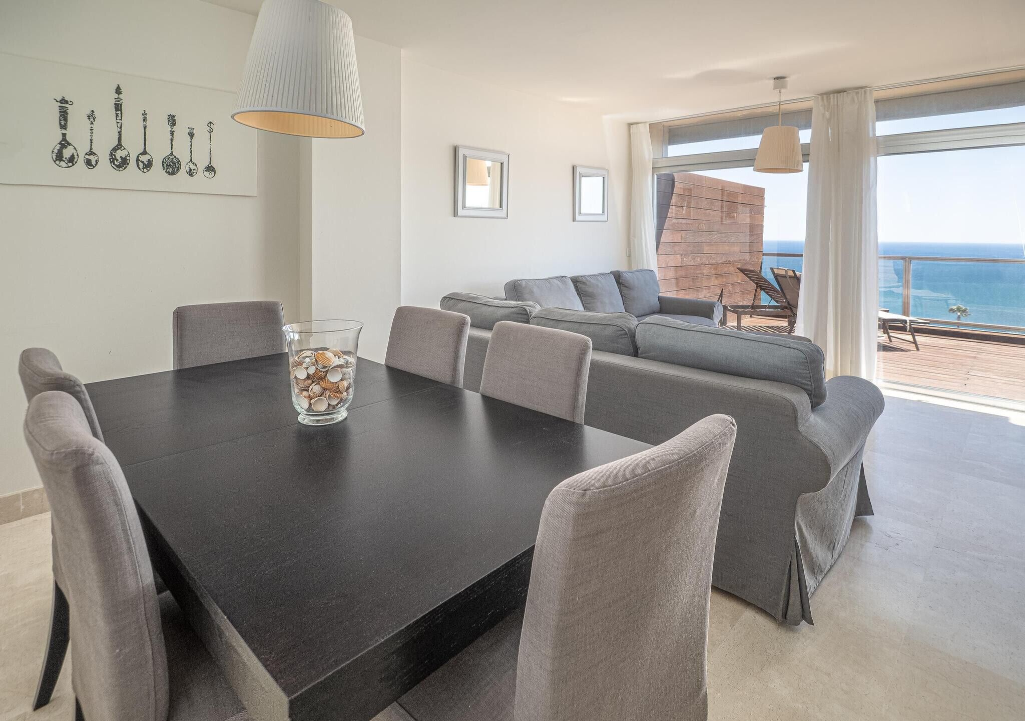 Un salon moderne avec une table à manger sombre, des canapés gris et un balcon offrant une vue sur la mer.