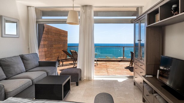 A modern living room with a sofa, a coffee table, and a TV on a wooden cabinet. There is a balcony with a view of the ocean. - Punta Paloma Penthouse 2364 Sea View (Manilva)