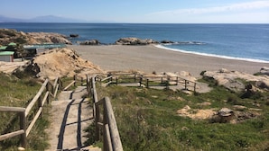 A coastal beach with a wooden staircase leading down to the shore, a small building with a green roof, and rocky terrain. - Punta Paloma Penthouse 2364 Sea View (Manilva)