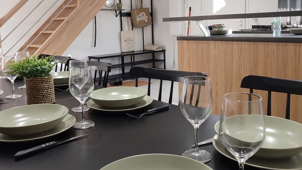 Une salle à manger avec une table noire dressée pour quatre personnes, comprenant des assiettes, des verres et des couverts. Un escalier avec des rampes en bois est visible à l’arrière-plan.