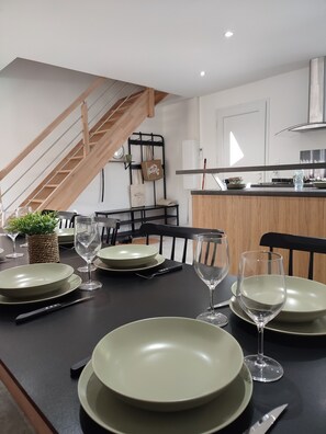 A dining area with a black table set for four, including plates, glasses, and cutlery. A staircase with wooden railings is visible in the background. - HOUSE BY THE SEA (Agde)