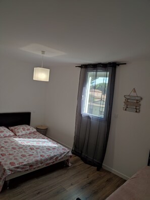A bedroom with a bed, a hanging light fixture, a window with curtains, and a wall-mounted sign. - HOUSE BY THE SEA (Agde)
