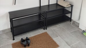 Un banc en métal noir, sur lequel est posé un tapis tissé, et une paire de chaussures noires sur le sol. Une petite étagère supporte une plante et un miroir. Un sac suspendu, sur lequel on peut lire le mot « Sabaotte », est visible.