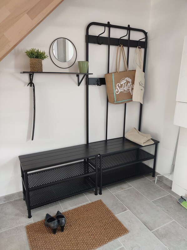 A black metal bench with a woven rug and black shoes on the floor. A small shelf holds a plant and a mirror. A hanging bag with "Sabaotte" text is visible. - HOUSE BY THE SEA (Agde)