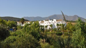 A white building with multiple floors, surrounded by greenery and mountains in the background.