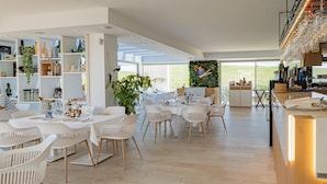 A spacious dining area with white tables and chairs, a large plant, and a view of the outdoors.