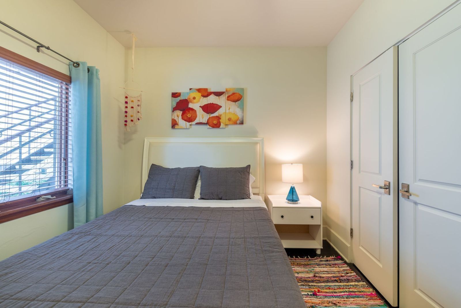 Een slaapkamer met een bed, nachtkastje, lamp, kleurenbekend tapijt, een deur en een raam met jaloezieën.