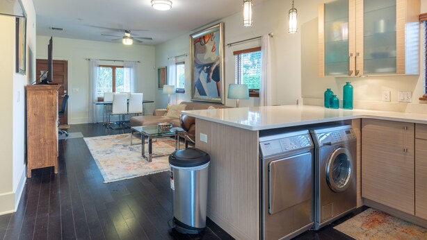 A modern kitchen with a built-in washer and dryer, a dining area with a glass table, and a living room with a large painting. - Light, Bright and Full of Delight! 2 Blocks to Park Ave (Winter Park)