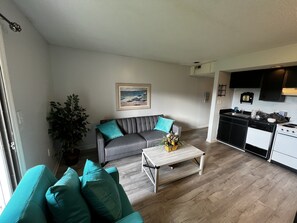 Relax in this cozy space with a sofa, coffee table, and kitchenette, surrounded by a potted plant and a beautiful landscape painting.
