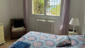A bedroom with a bed, a radiator, a wicker chair, a nightstand with a lamp, and a view of greenery and water through a window. - BEAUTIFUL 3-ROOM APARTMENT WITH SEA VIEW - LES ISSAMBRES (Roquebrune-sur-Argens)