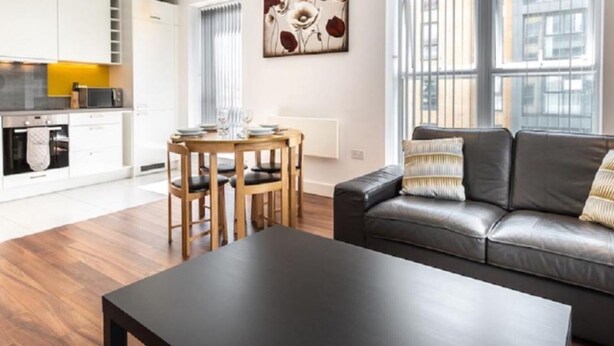 A modern living room with a black leather sofa, a wooden dining table with chairs, and a kitchen area with white cabinets and appliances. - Impeccable 2-bed Apartment in Reading (Reading)