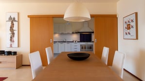 A modern dining area with a wooden table and white chairs, a kitchen in the background, and artwork on the walls. - Apartment (Marina di Ragusa)